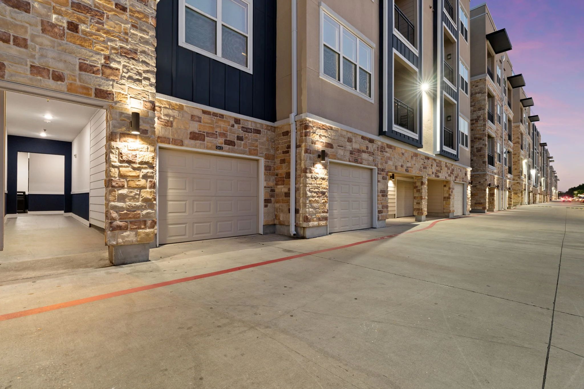 An exterior view of apartment buildings featuring attached garages, stone siding, and a paved driveway at dusk.